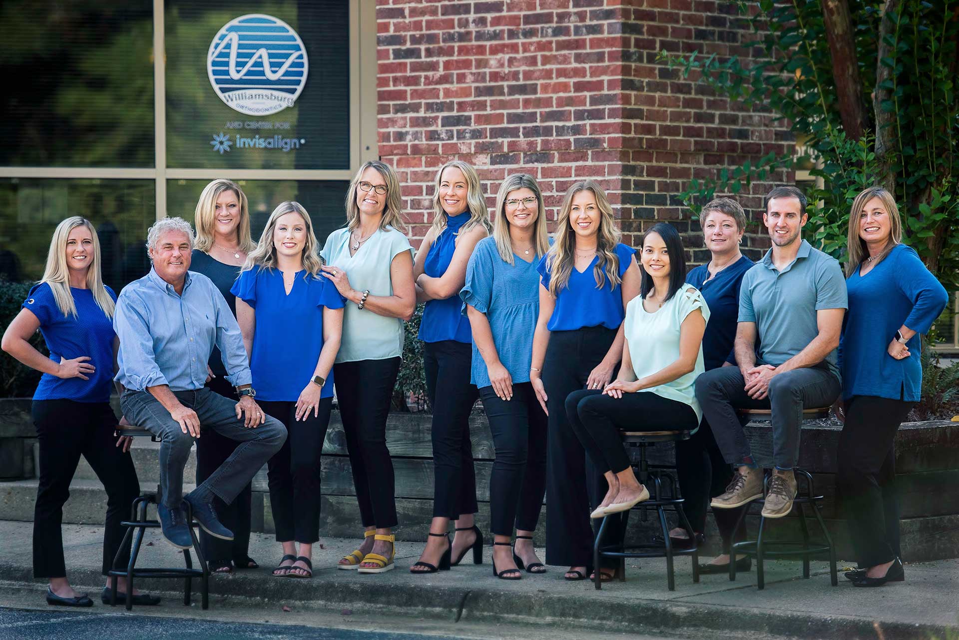 Group photo of the Williamsburg Orthodontics team standing and sitting together outside the office in front of a brick wall.
